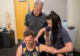 A student adjusting a rugby player's shoulder as a chiropractic instructor looks on.