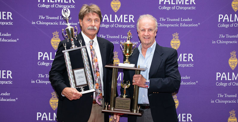 Two men holding rugby trophies in front of purple Palmer backdrop.