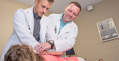 Instructor guiding student's hand positioning on a patient's upper back.
