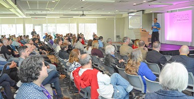 Speaker with microphone in front of large seated audience.