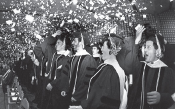 Black and white photo of students in graduation gowns throwing cards into the air.