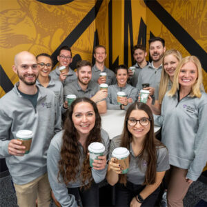 group of students with coffees in front of gold wall.