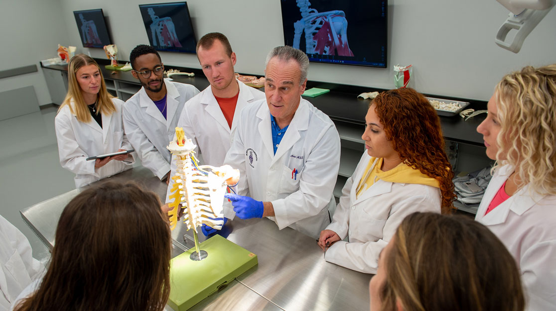 Students in white lab coats gathered around instructor using spine model.