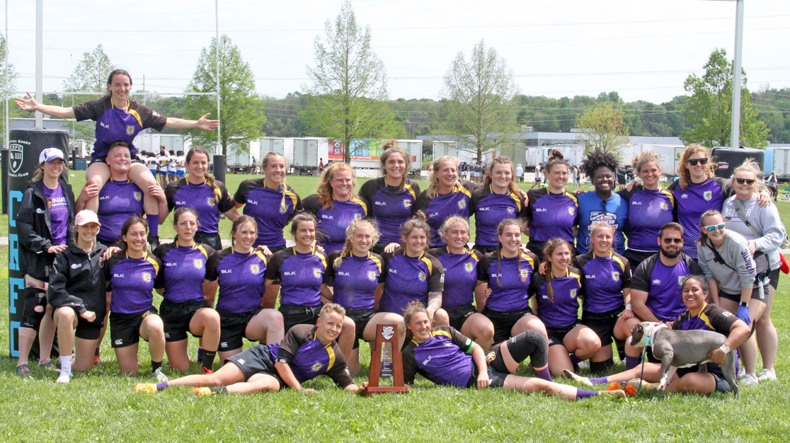 Group of women's rugby players in purple uniforms smiling outside with trophy.