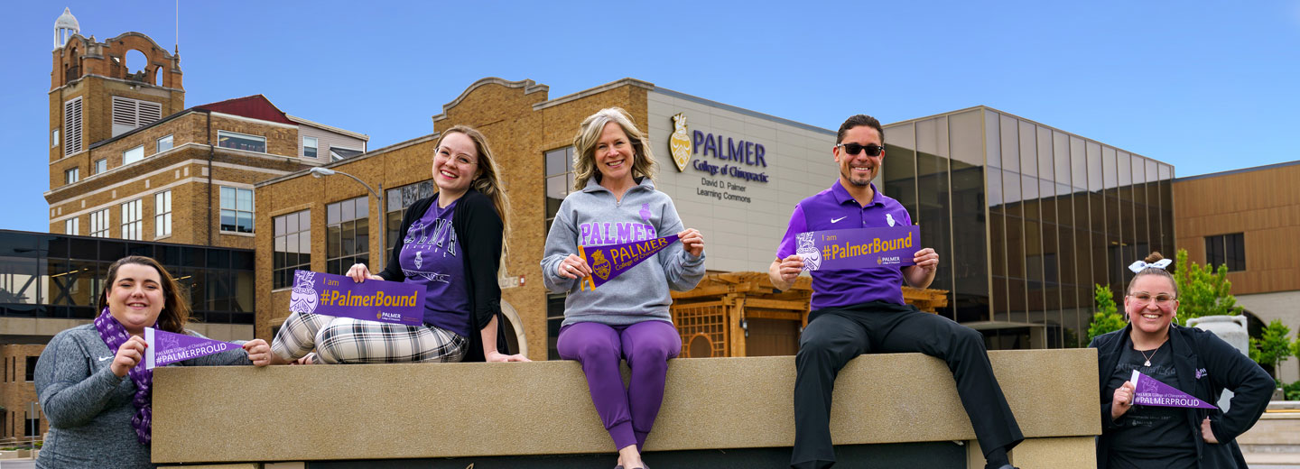 Five counselors in purple sitting in front of Palmer College building holding Palmer Bound pennants.
