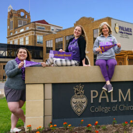 Three counselors in purple sitting in front of Palmer College building holding Palmer Bound pennants.