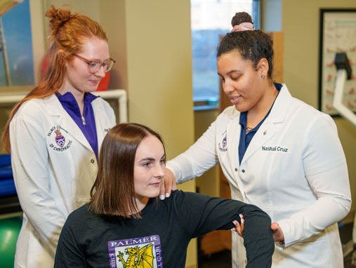 Two students in white coats evaluating patient's shoulder movement.