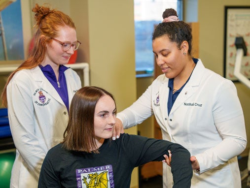 Two students in white coats evaluating patient's shoulder movement.