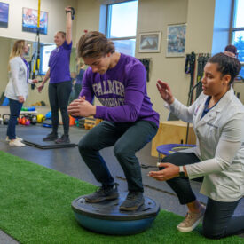 Sports rehab students working with a patient lifting a weight and a patient balancing on rehab equipment.