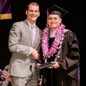 Student in graduation gown and lei accepting award and shaking hands with professor.