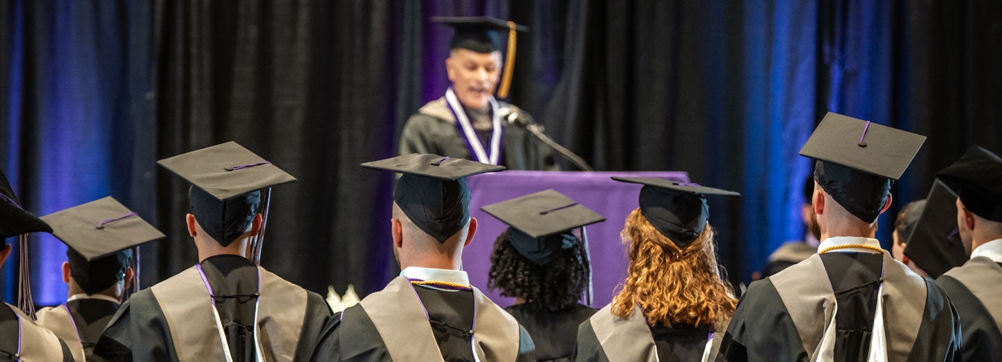 Students in commencement cap and gowns listening to speaker.