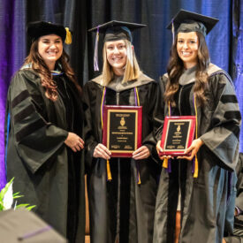 Three women in commencement caps and gowns holding awards.