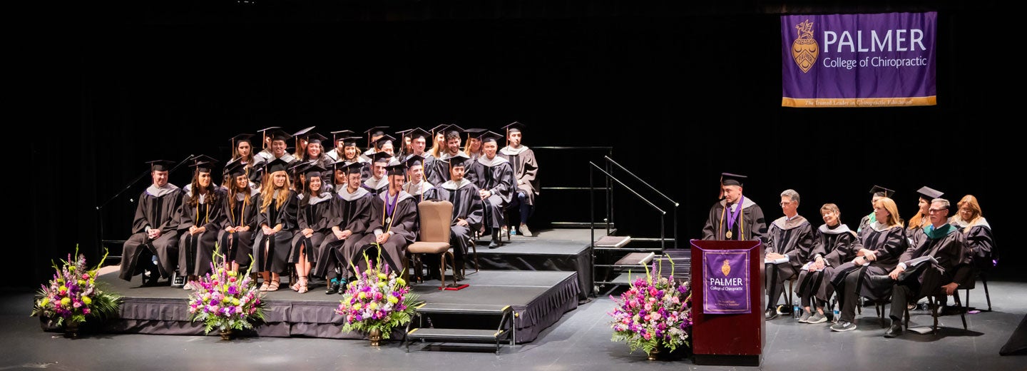 Speaker at podium in front of sitting graduates in cap and gowns.