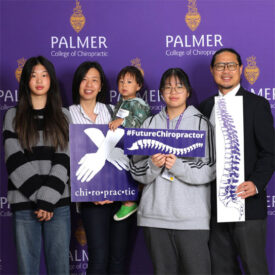 Family posing in front of purple Palmer College backdrop holding signs.