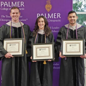 Three students in graduation cap and gowns holding awards.