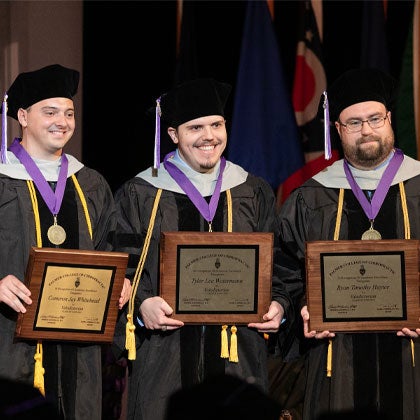 Three students in graduation cap and gowns holding awards.