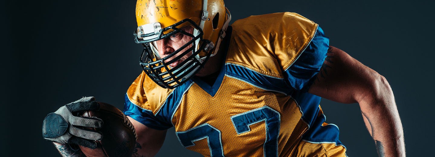 Football player in gear holding a football against a black background.